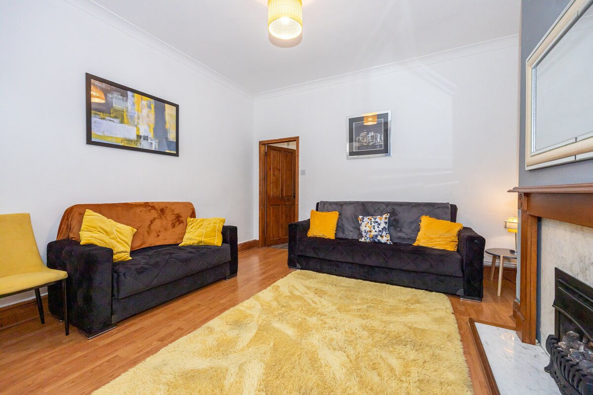 The living room features two dark sofas, complemented by a yellow chair. A plush area rug covers the wooden floor, and artwork adorns the walls. Soft lighting creates a welcoming ambience, while a fireplace adds a touch of warmth to the space.
