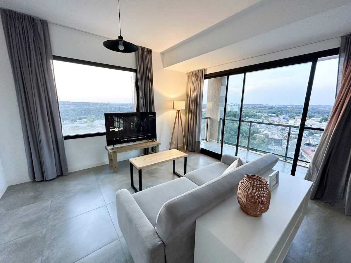 A modern living area is presented with large windows offering panoramic city views. A light-colored sofa faces a wooden coffee table and a television, while stylish floor lamps and grey curtains complement the neutral décor. The space is filled with natural light.