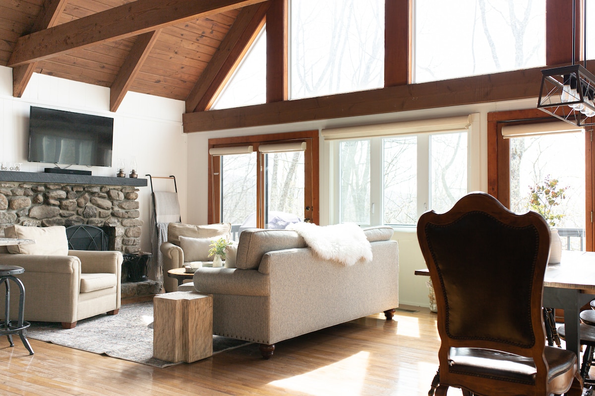 The living area features a stone fireplace and large windows that allow natural light to flood the space. Soft sofas are arranged around a wooden coffee table, complemented by a dining area with a classic wooden chair. Neutral tones enhance the warm ambiance.