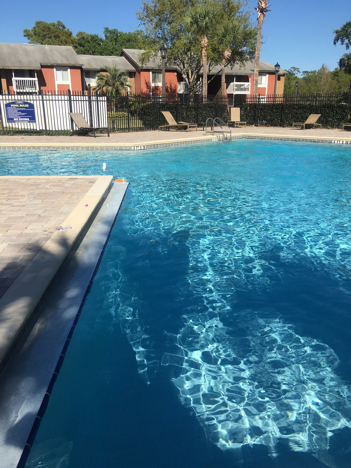 A clear blue pool reflects sunlight, with a pool deck featuring lounge chairs positioned around the perimeter. Lush green trees and well-maintained landscaping are visible in the background, enhancing the serenity of the outdoor setting.