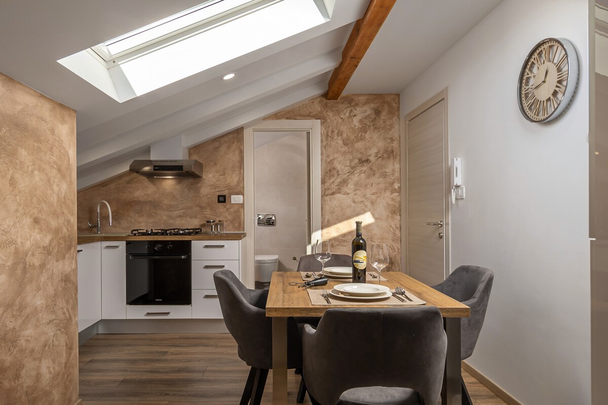 A modern kitchen and dining area are highlighted, featuring a wooden dining table set for two. Grey upholstered chairs surround the table. Natural light enters through a skylight, illuminating the sleek cabinetry and contemporary appliances in the kitchen.