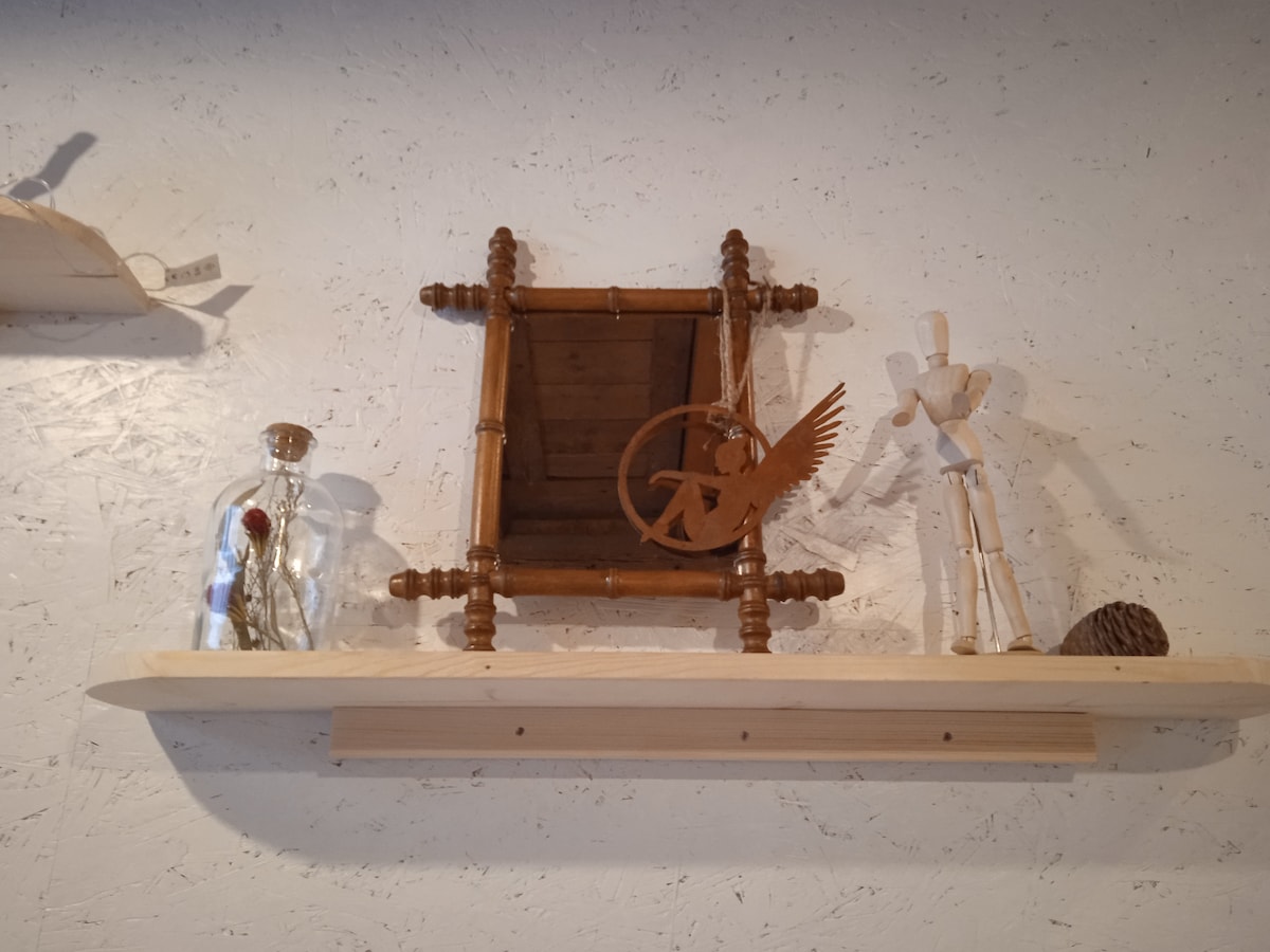 A wooden shelf displays a framed artwork featuring a bird and a stylized figure made of wood. An elegant glass bottle containing a single rose is positioned next to these items, adding a touch of decor. A small pine cone is also present on the shelf.