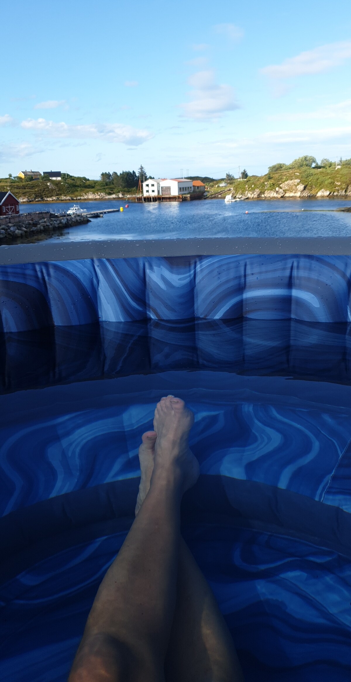 A view from a hot tub, featuring feet submerged in water. The background displays a serene landscape with a calm body of water, dotted with boats and surrounded by lush greenery. Distant structures are visible, reflecting the natural setting.