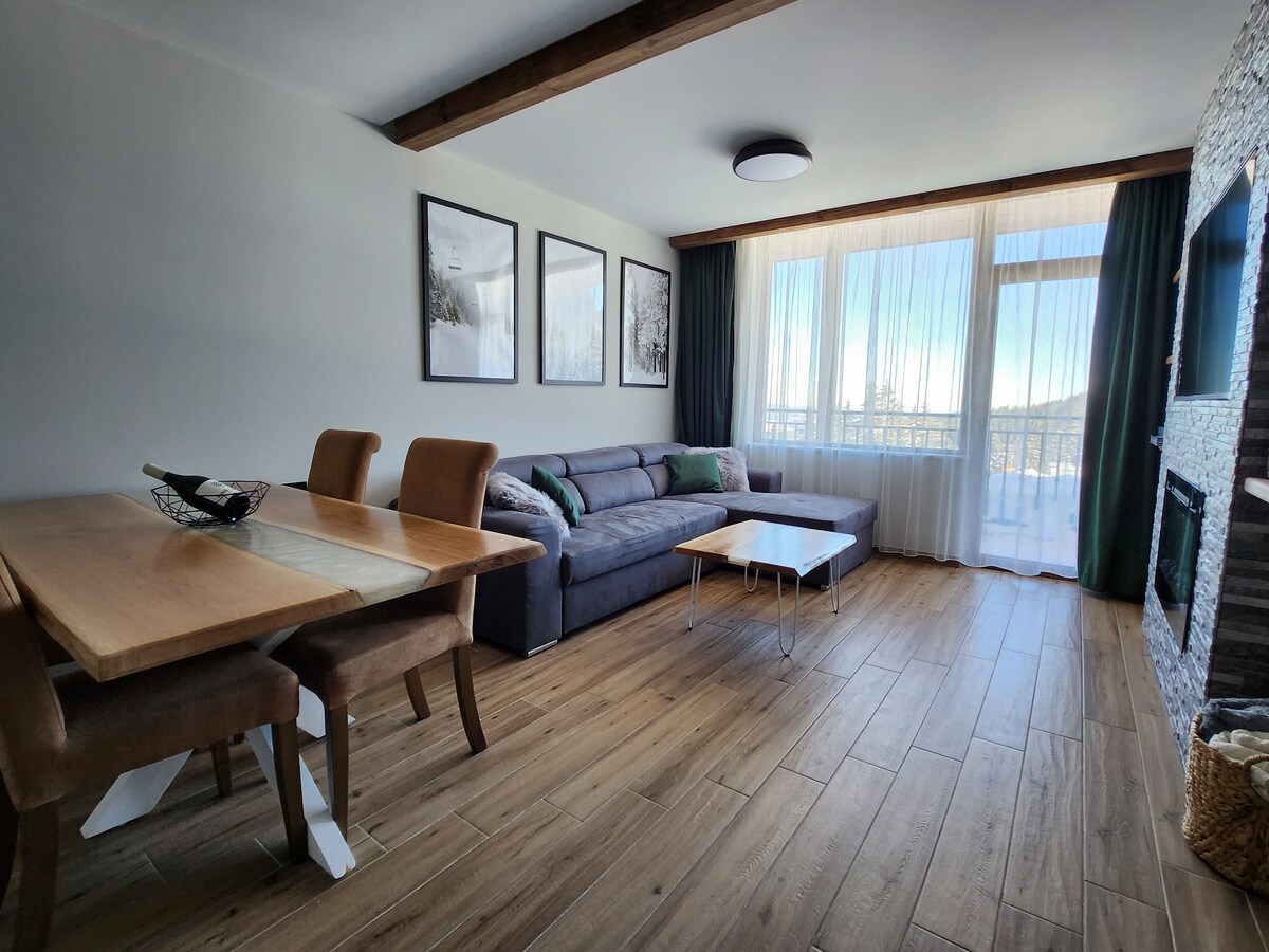A spacious living area features a comfortable gray sofa paired with a wooden coffee table. A dining table with four chairs is present, complemented by large windows that allow natural light to fill the room. Scenic mountain views are framed by sheer curtains.