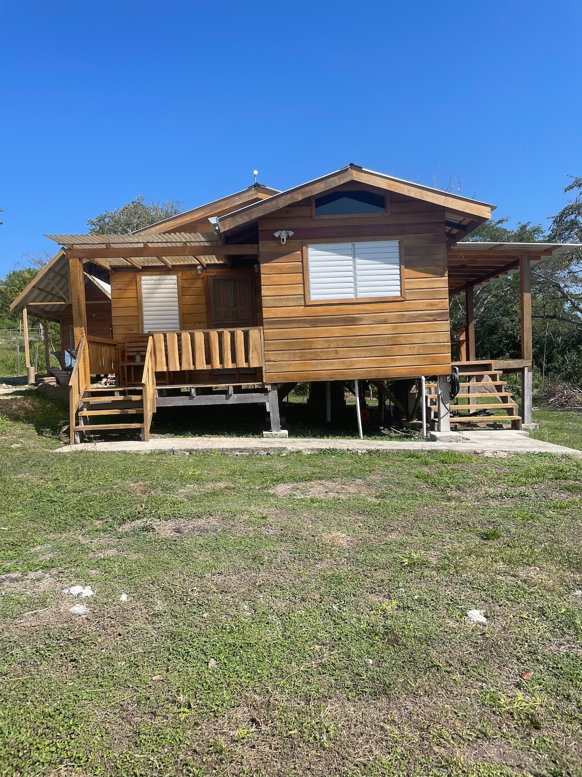 A cozy wooden cabin is set on stilts, featuring a welcoming front porch with steps leading up. The exterior showcases warm wood tones against a clear blue sky. Grass surrounds the cabin, emphasizing a natural atmosphere.