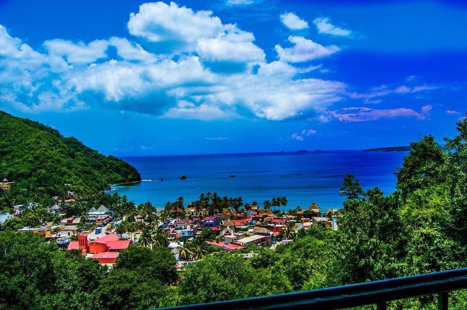 A panoramic view of a vibrant coastal town is captured, showcasing colorful buildings nestled among lush greenery. Gentle waves lap at the shore under a bright blue sky, dotted with clouds. The surrounding landscape features a combination of hills and the expansive ocean.