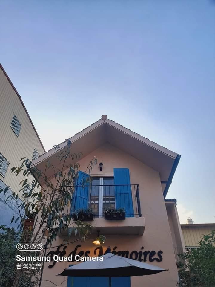 A charming two-story structure features a peaked roof and blue shutters. A small balcony is adorned with a railing and a flower pot. The building's name is displayed prominently on the lower wall, complemented by surrounding greenery.