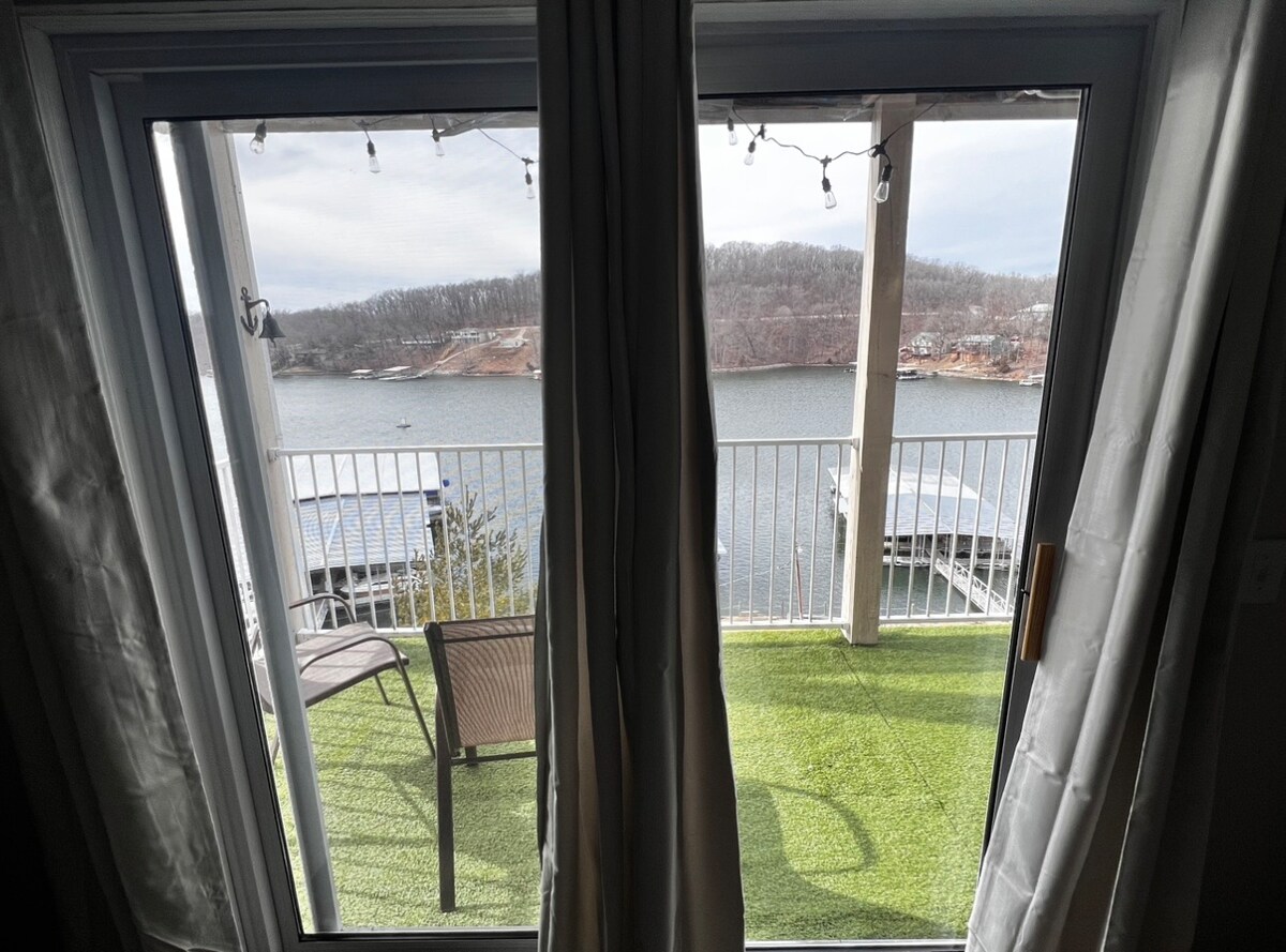 A sliding glass door provides access to a private balcony overlooking a tranquil lake. Outdoor seating is visible on the balcony, with green artificial turf underfoot. Soft string lights are hung along the railing, and the serene water reflects the surrounding landscape.