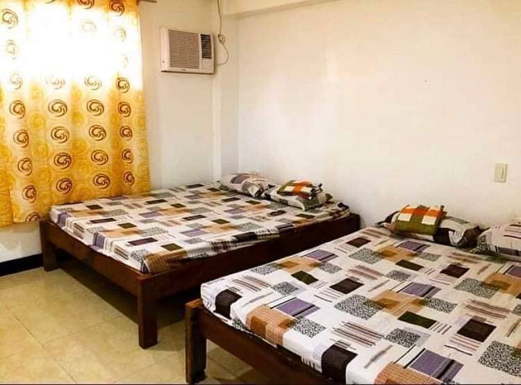 An air-conditioned bedroom features two queen-sized beds with patterned beddings. Natural light enters through a window covered by bright, patterned curtains, illuminating the light-colored tiled floor. A wall-mounted air conditioning unit is positioned above the window.
