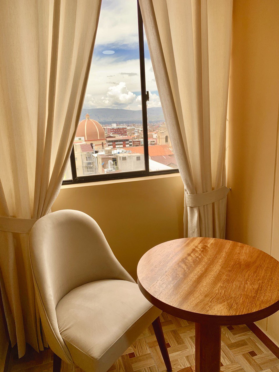 A small round wooden table sits beside a plush chair, positioned near a window. Soft, neutral curtains frame the window, allowing natural light to illuminate the space. A view of the city skyline and the distant dome is visible beyond the glass.