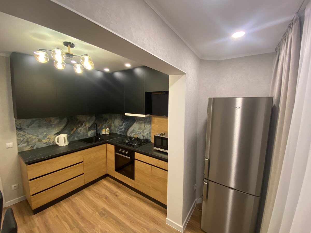 A modern kitchen is presented, featuring dark cabinetry and light wooden accents. Countertops are made of dark material, with a marble backsplash adding color. A stainless steel refrigerator stands nearby, complemented by a gas stove and microwave, all illuminated by warm lighting fixtures.