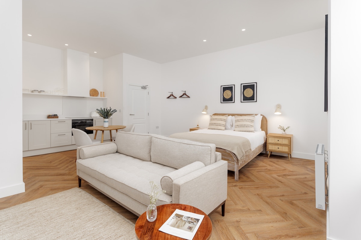 A spacious studio apartment is showcased, featuring a cozy lounge area with a neutral sofa and a round wooden coffee table. The sleeping area includes a comfortable bed with soft bedding, complemented by nightstands and artwork on the walls. A modern kitchen is visible in the background.