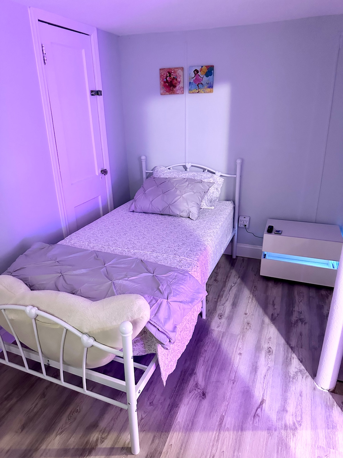 A small bedroom is highlighted with a white metal bed, dressed in light-colored bedding and additional pillows. A nightstand sits nearby, featuring subtle ambient lighting. Colorful wall art adds a touch of personality, while the floors are finished in a warm wood tone.