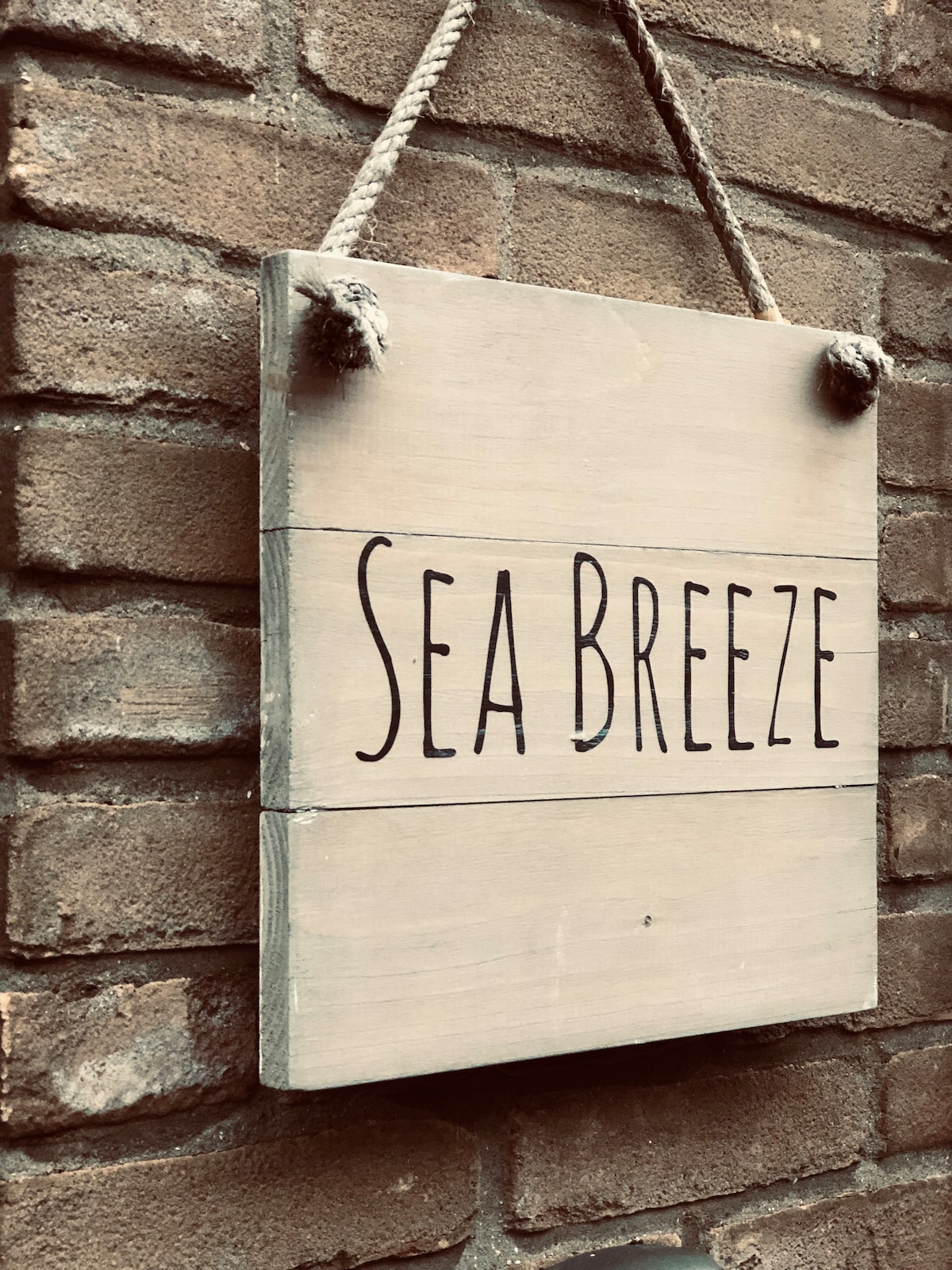A wooden sign hanging from a rope is attached to a brick wall. The sign features the text 'Sea Breeze' in simple, black lettering, presenting a welcoming touch to the entrance.