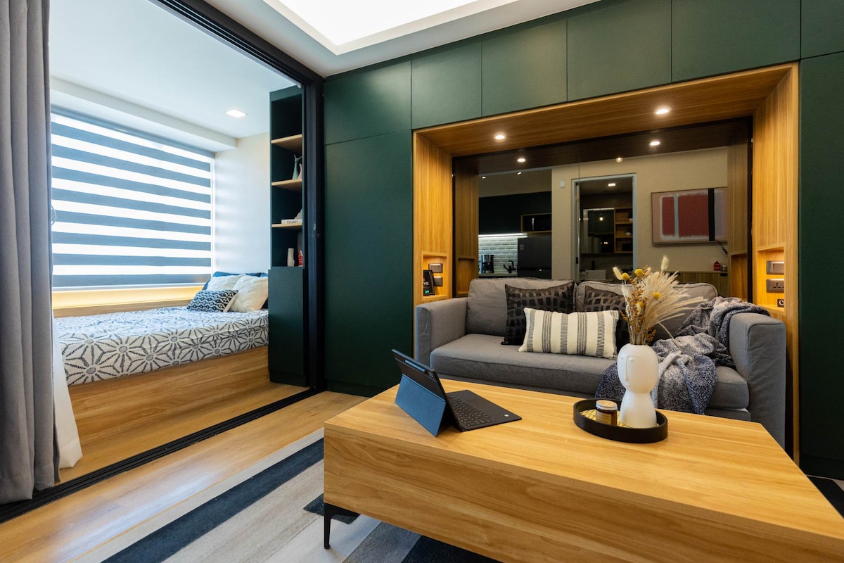 A spacious studio condo is shown, featuring a living area with a comfortable sofa and a wooden coffee table. A cozy sleeping area is visible through sliding doors, revealing a neatly made bed surrounded by natural light from window treatments.