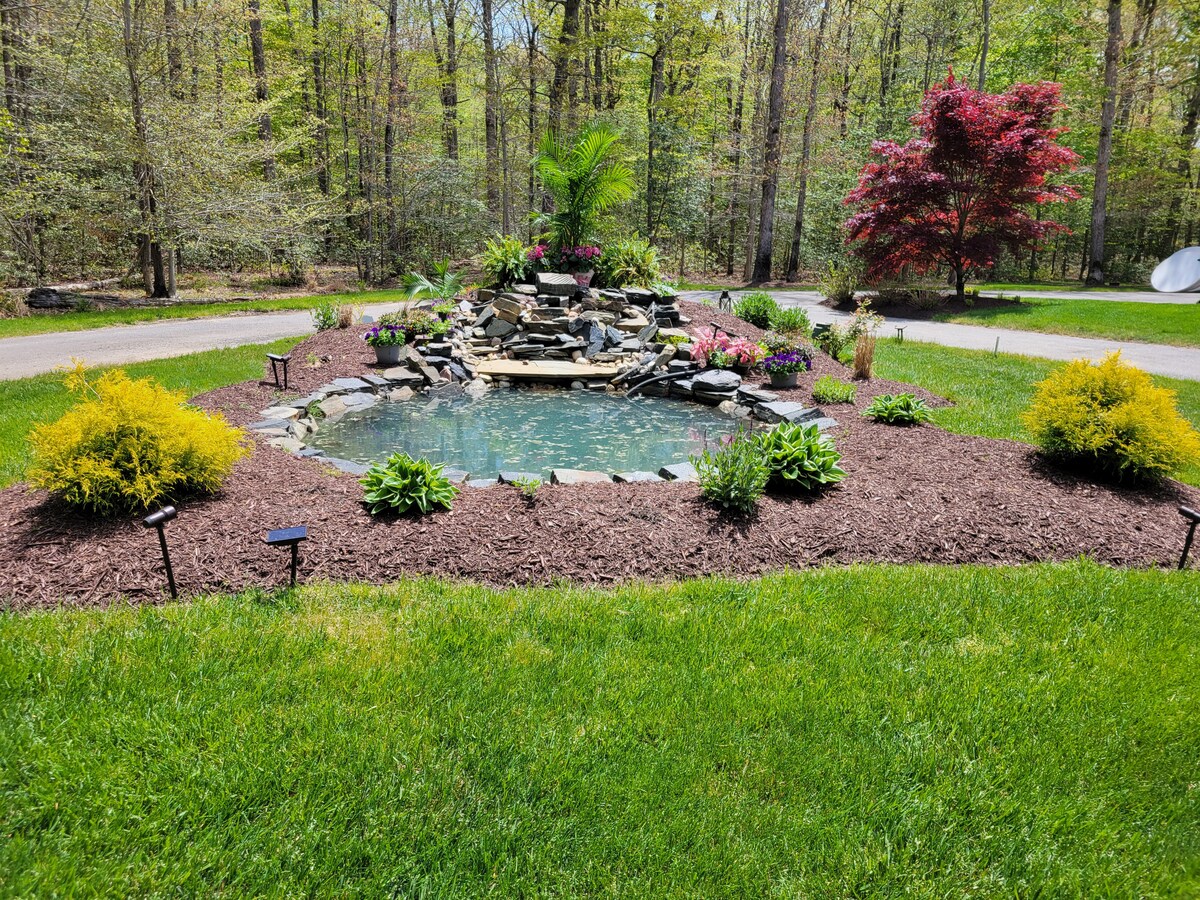 A serene pond is framed by lush green grass and vibrant landscaping, featuring various plants and flowers. A softly flowing waterfall cascades over rocks, creating a calming focal point. Natural light filters through the surrounding trees, enhancing the tranquil setting.