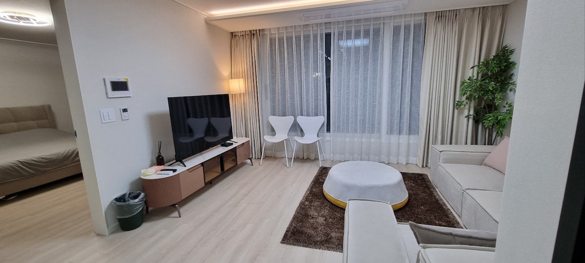 A bright living area features a white sofa and two white chairs positioned around a soft circular rug. A television is mounted on the wall, and large windows are draped with sheer curtains, allowing natural light to fill the space.