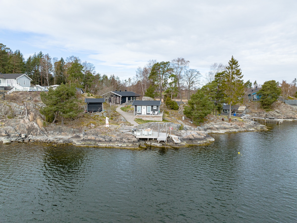 A serene waterfront view showcases a collection of cozy buildings nestled among trees. Two private docks extend into the calm waters, providing easy access for swimming. The surrounding landscape features rocky terrain and lush greenery, creating a peaceful outdoor setting.