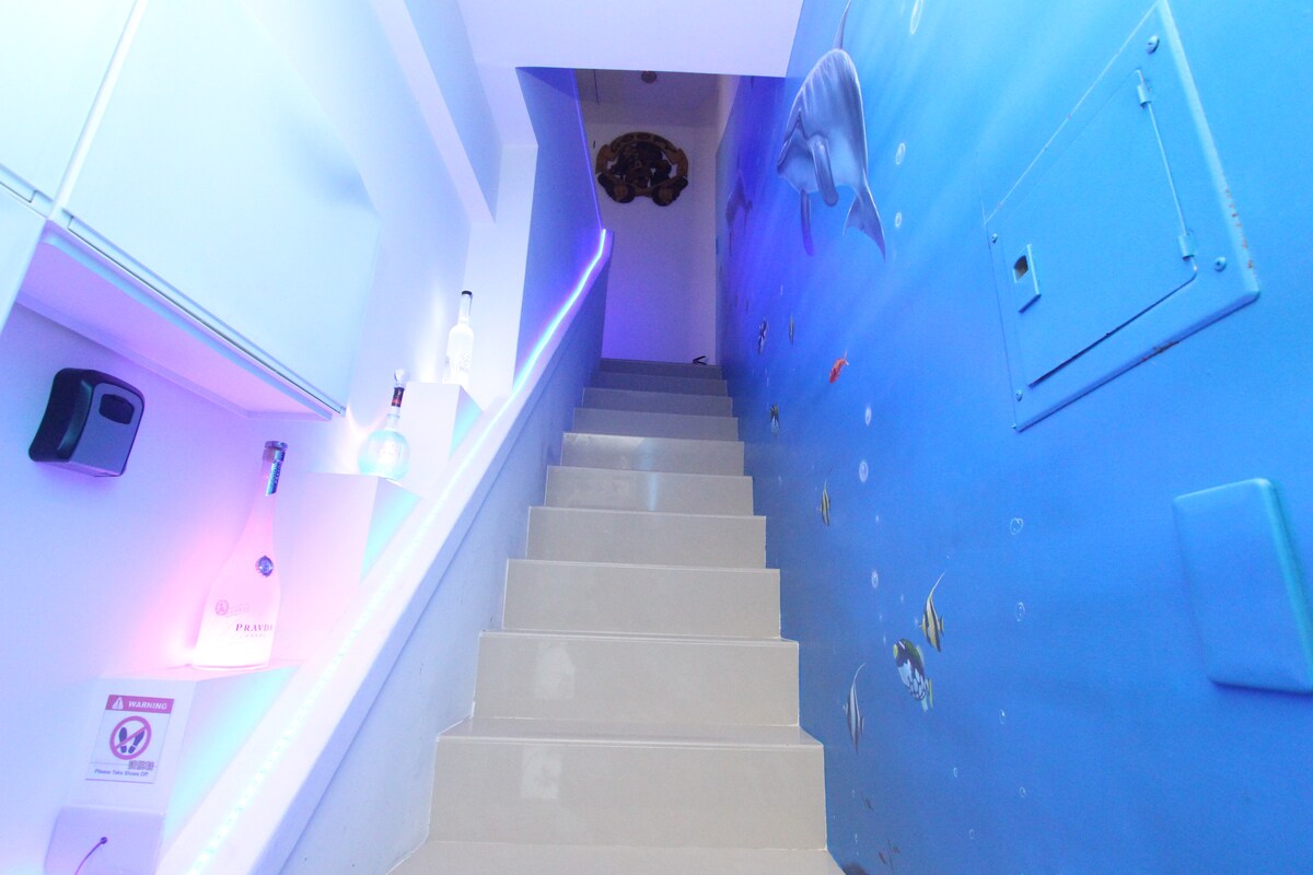 A bright stairway leads upward, adorned with ocean-themed murals depicting various marine life. Soft purple lighting outlines the steps, enhancing the modern ambiance. The walls are painted in soothing blue, creating a serene and inviting transition to the upper level.