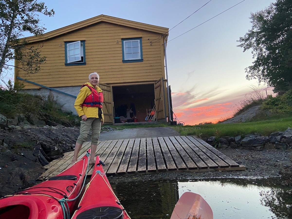 A yellow house with blue trim is viewed from the perspective of two red kayaks, positioned near a wooden ramp leading to the water. The evening sky displays soft hues of orange and pink as the sun sets behind the house.