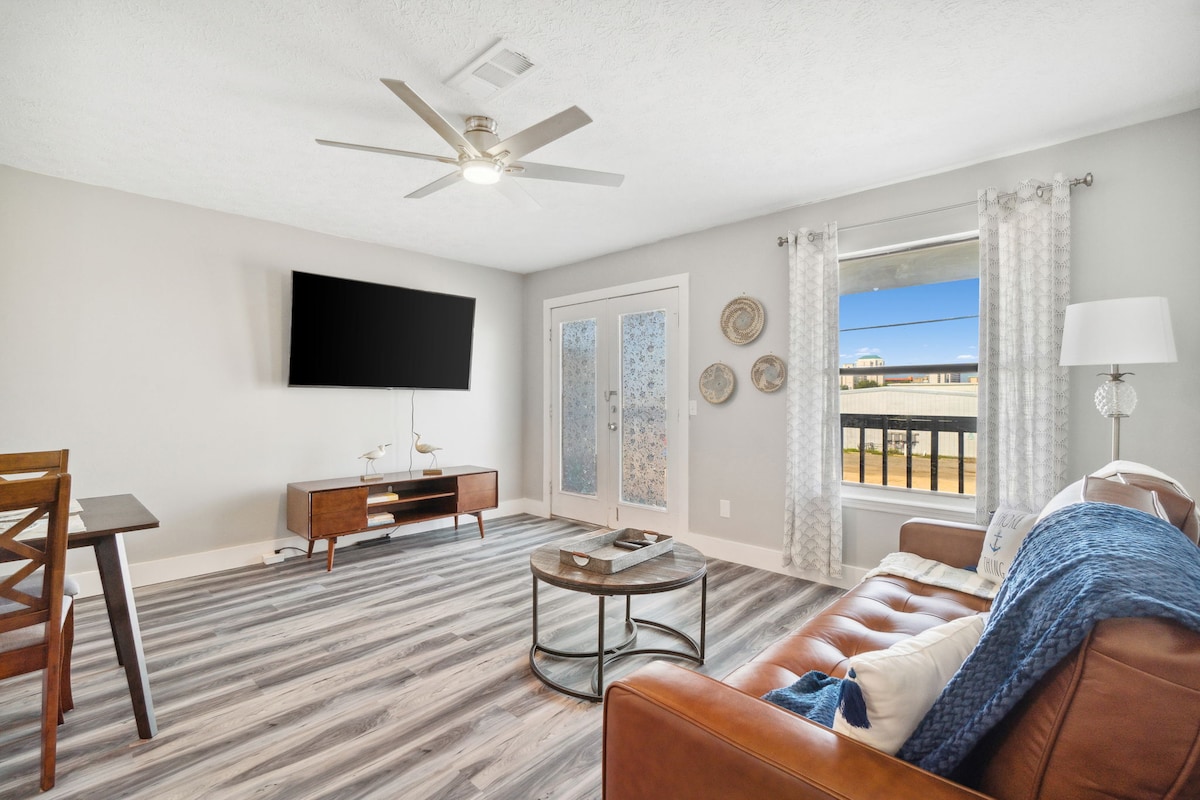A living area featuring a brown leather sofa adorned with a blue throw, a modern round coffee table, and a wooden dining table. Natural light enters through a glass door leading to a balcony, while a ceiling fan and wall-mounted television complete the space.