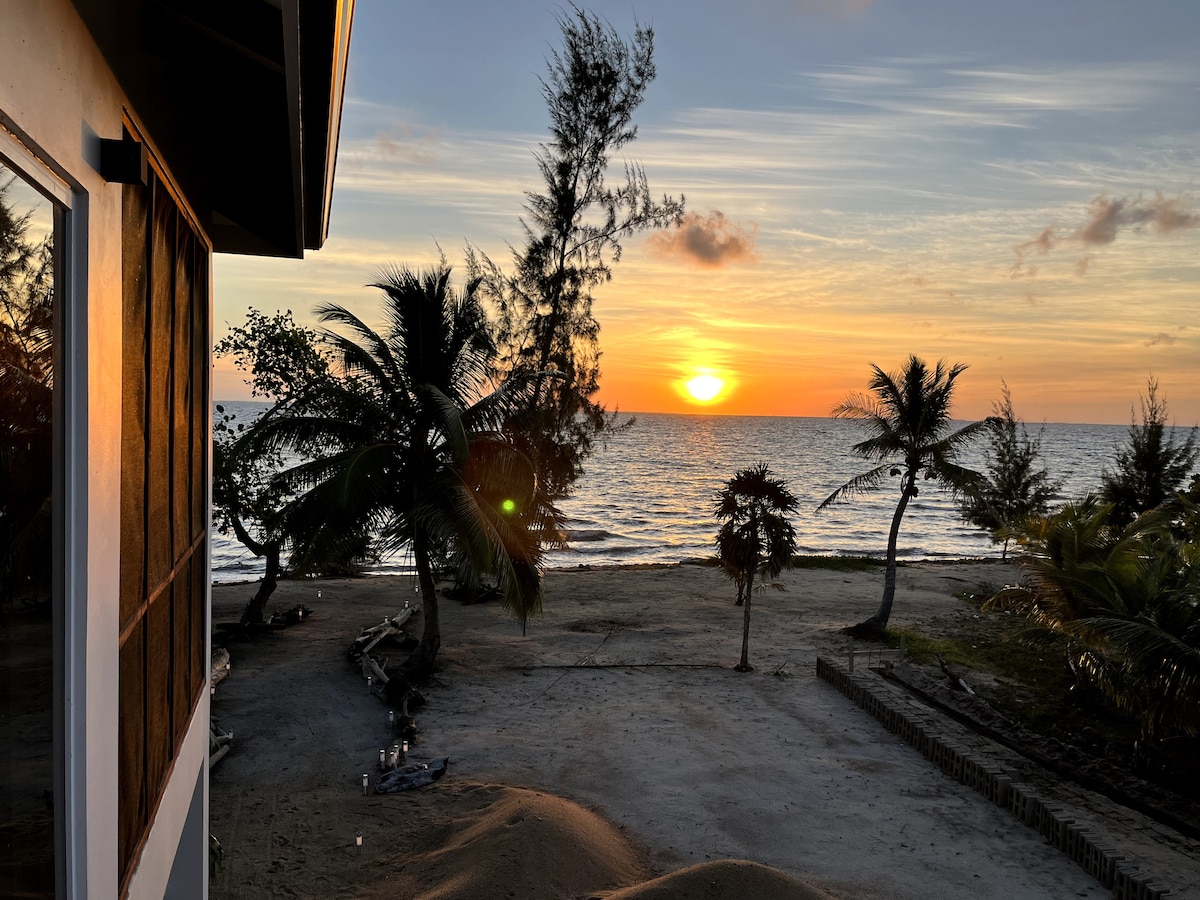 A serene sunrise is captured over the Caribbean Ocean, with sunlight reflecting on the water's surface. Silhouettes of palm trees and distant vegetation frame the scene. The sandy beach extends toward the horizon, providing a tranquil environment for relaxation.
