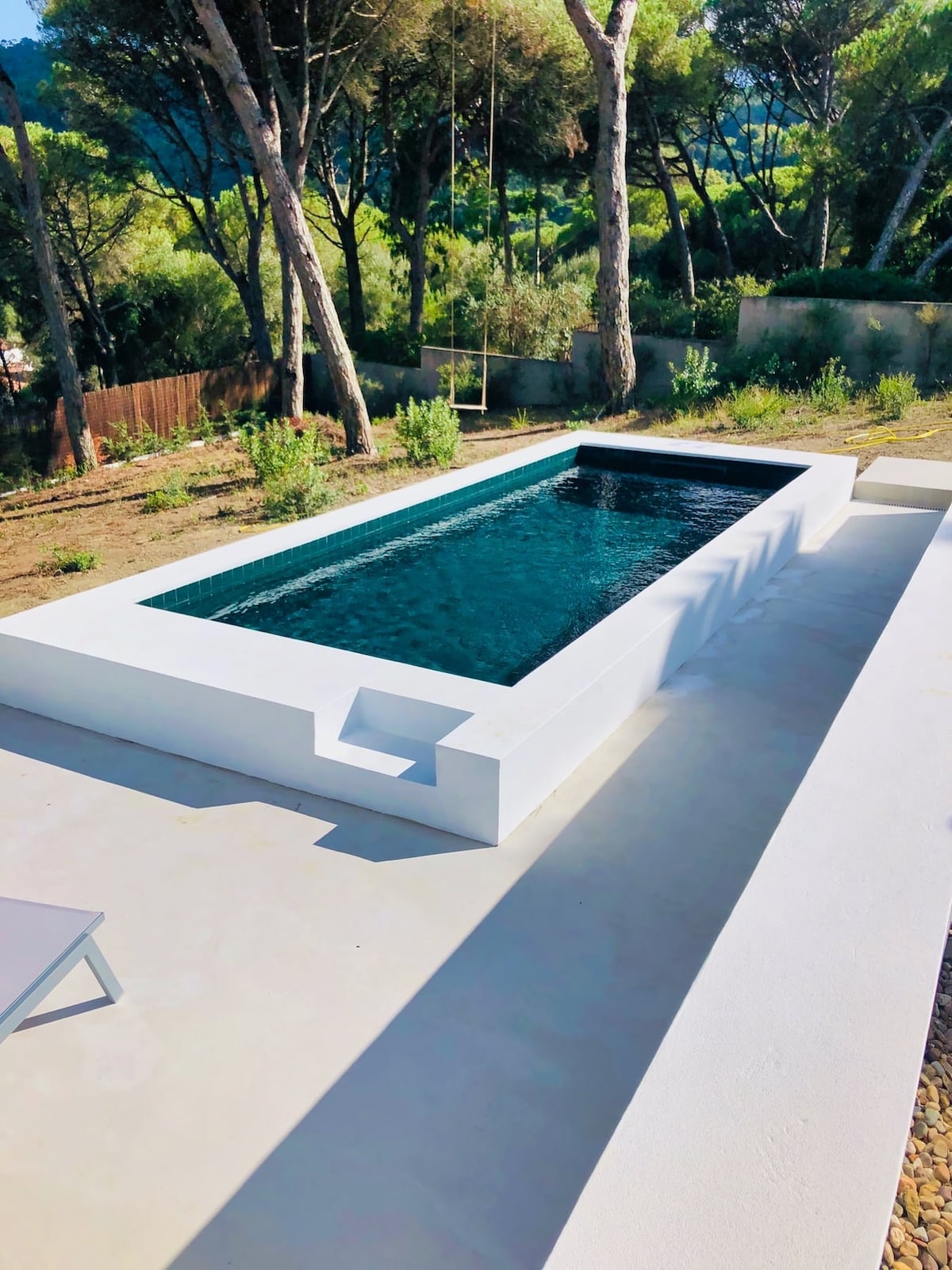 A modern pool is set on a smooth surface, surrounded by greenery and tall trees. The pool features a minimalist design with clean lines, reflecting the sky's blue hues. A lounge chair is positioned nearby, inviting relaxation.