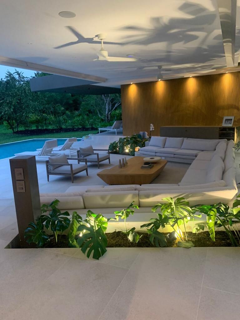 An expansive outdoor seating area features a large, curved sectional sofa surrounding a wooden coffee table. Lush greenery is noticed in the planter areas, with modern lounge chairs positioned nearby, all set against a backdrop of a serene pool illuminated in soft lighting.
