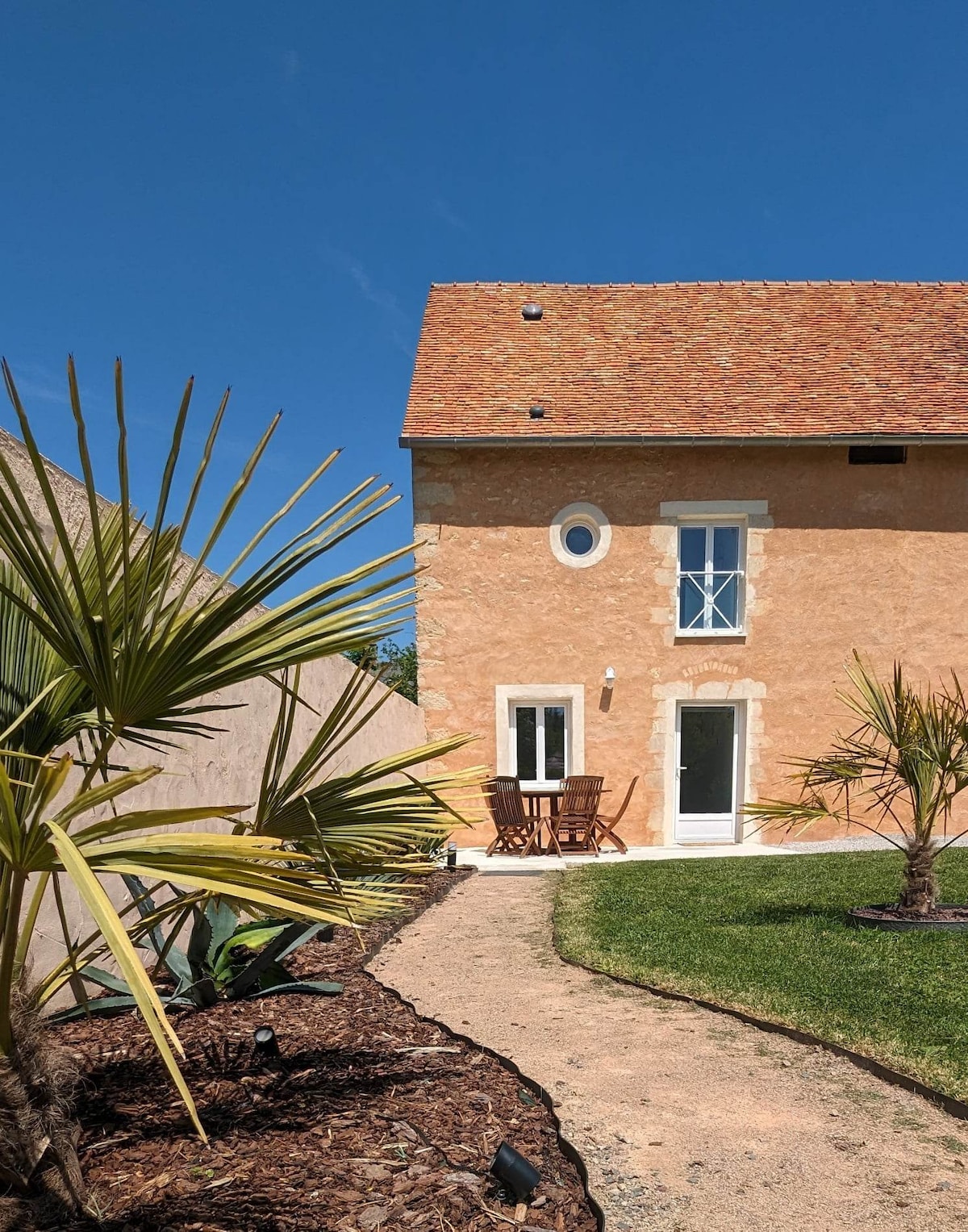 The exterior of the house is highlighted by a warm-toned façade and a red-tiled roof. A small wooden table with chairs is positioned near a secluded door. Palm trees line the pathway, which leads to a well-maintained grassy area under a clear blue sky.