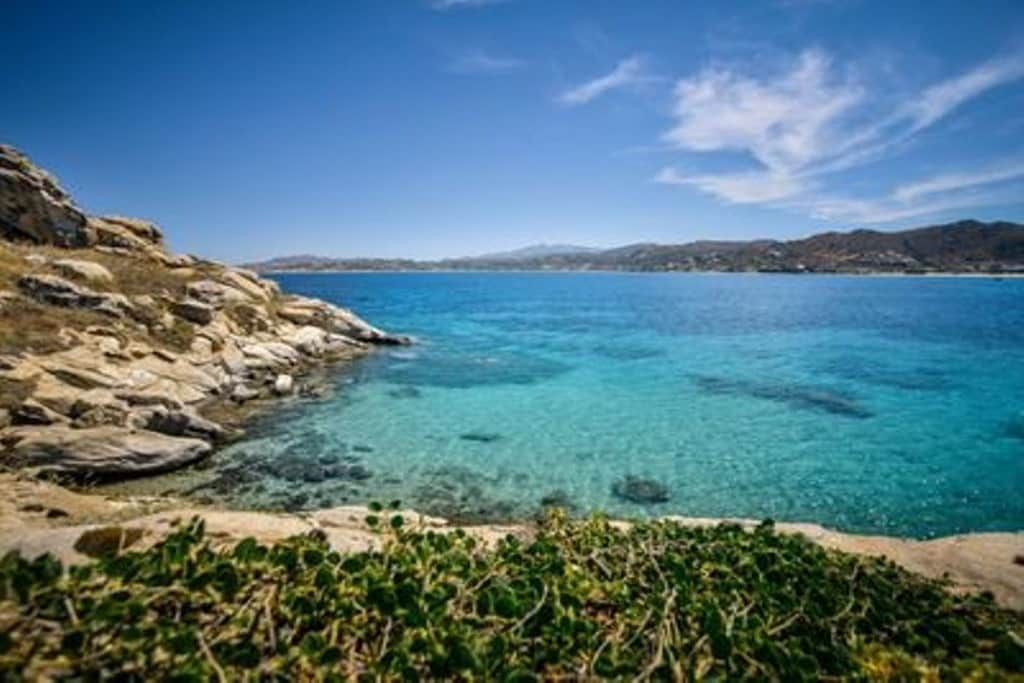 Clear turquoise waters are seen lapping gently against rocky shores, with a hint of vibrant green vegetation in the foreground. The horizon features distant hills and a blue sky embellished with wispy clouds, creating a serene coastal backdrop.