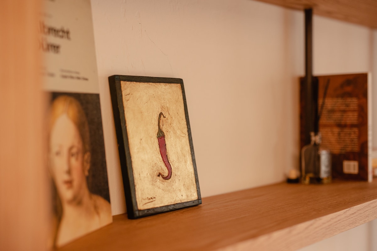A wooden shelf displays a selection of books and a framed artwork featuring a purple chili pepper. The shelf's natural material complements the warm tones of the decor, creating an inviting focal point in the space.