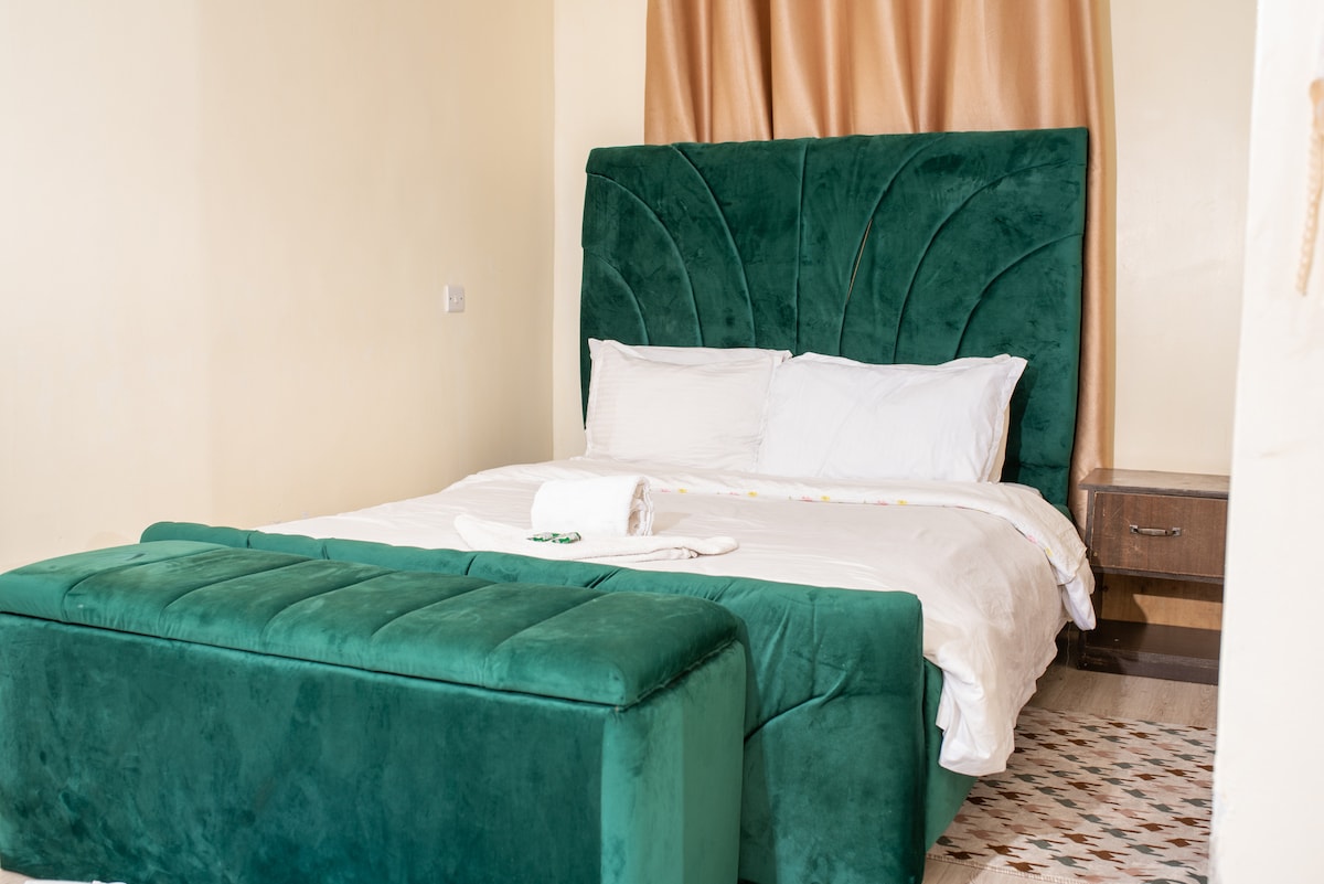 A spacious bedroom is highlighted by a large green upholstered headboard, framing a neatly made bed with crisp white linens. A coordinating bench sits at the foot of the bed. Natural light fills the room, complemented by soft, neutral wall tones.