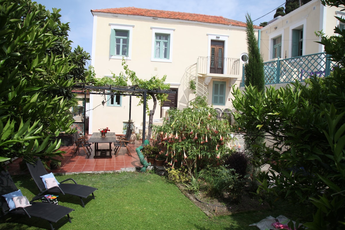 A garden area features a lush green lawn surrounded by vibrant plants. A patio with a wooden dining table is visible, complemented by two lounge chairs. The two-story villa, adorned in a soft yellow hue, stands in the background, with blue window shutters adding a charming touch.