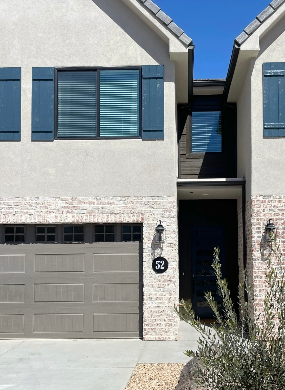 The exterior of a modern townhome is visible, featuring a combination of smooth grey walls and brick accents. Shuttered blue windows are situated above a large garage door, which is flanked by a light fixture. A manicured pathway leads to the front door, surrounded by minimal landscaping.