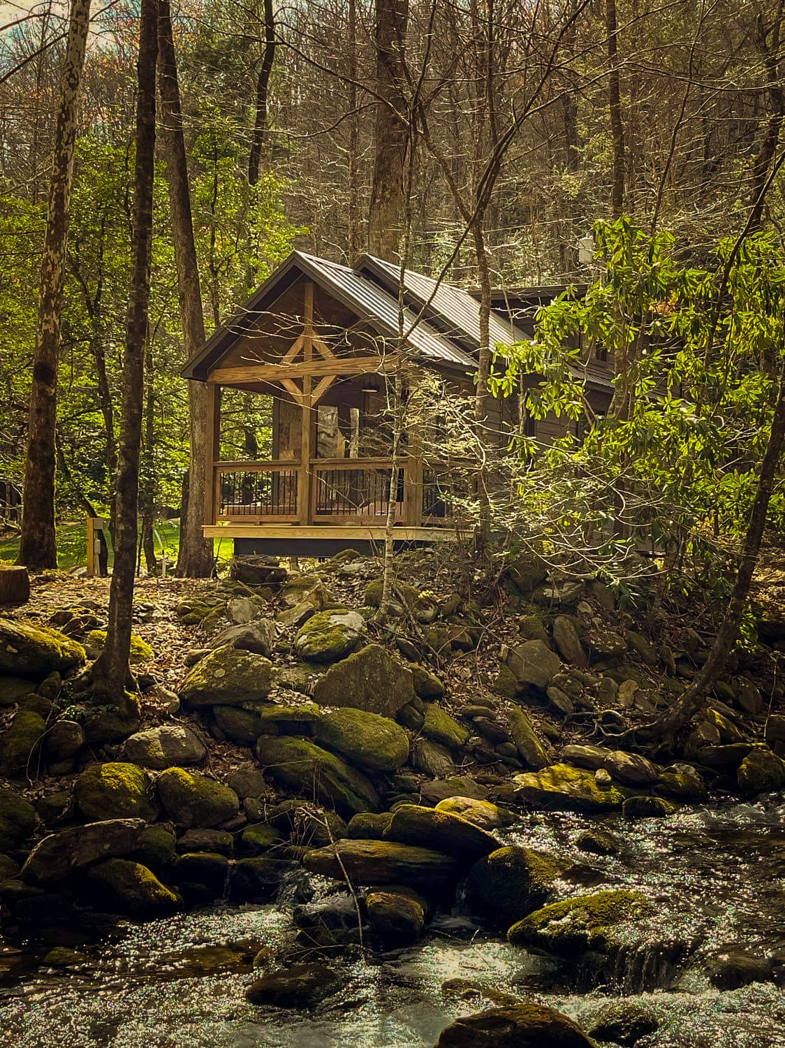 A cabin nestled among lush trees stands next to a gently flowing creek. The wooden structure features a triangular roof and expansive front porch. Smooth stones line the creek bank, complementing the natural landscape and creating a serene atmosphere.
