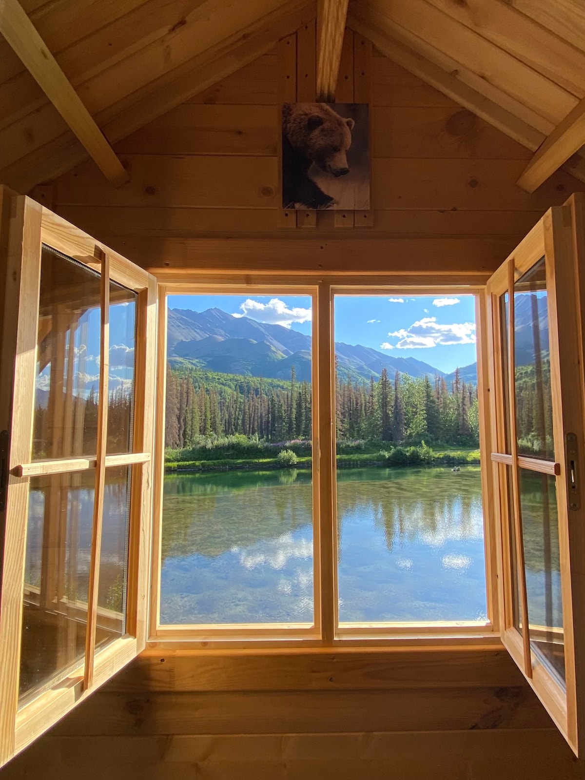 Large windows offer expansive views of the serene lake and surrounding mountains. The wooden interior is complemented by a mounted bear head, creating a rustic atmosphere. Natural light fills the space, highlighting the tranquil setting embraced by nature.