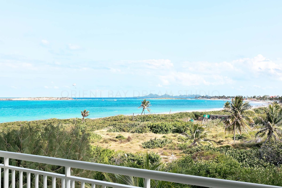 A panoramic view of the beach and sea is presented from the spacious balcony. Lush greenery frames the coastline, while gentle waves can be seen lapping at the shore. The sky is bright with scattered clouds, contributing to the serene atmosphere.