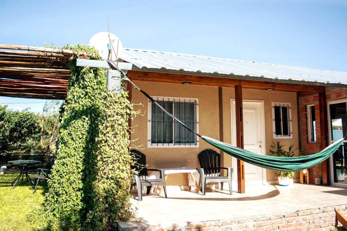 The exterior of a rural house features a spacious patio with a green hammock suspended between posts. A table and chairs are set for relaxation. Lush vegetation surrounds the area, providing a serene atmosphere. Large windows allow for ample natural light to enter the home.