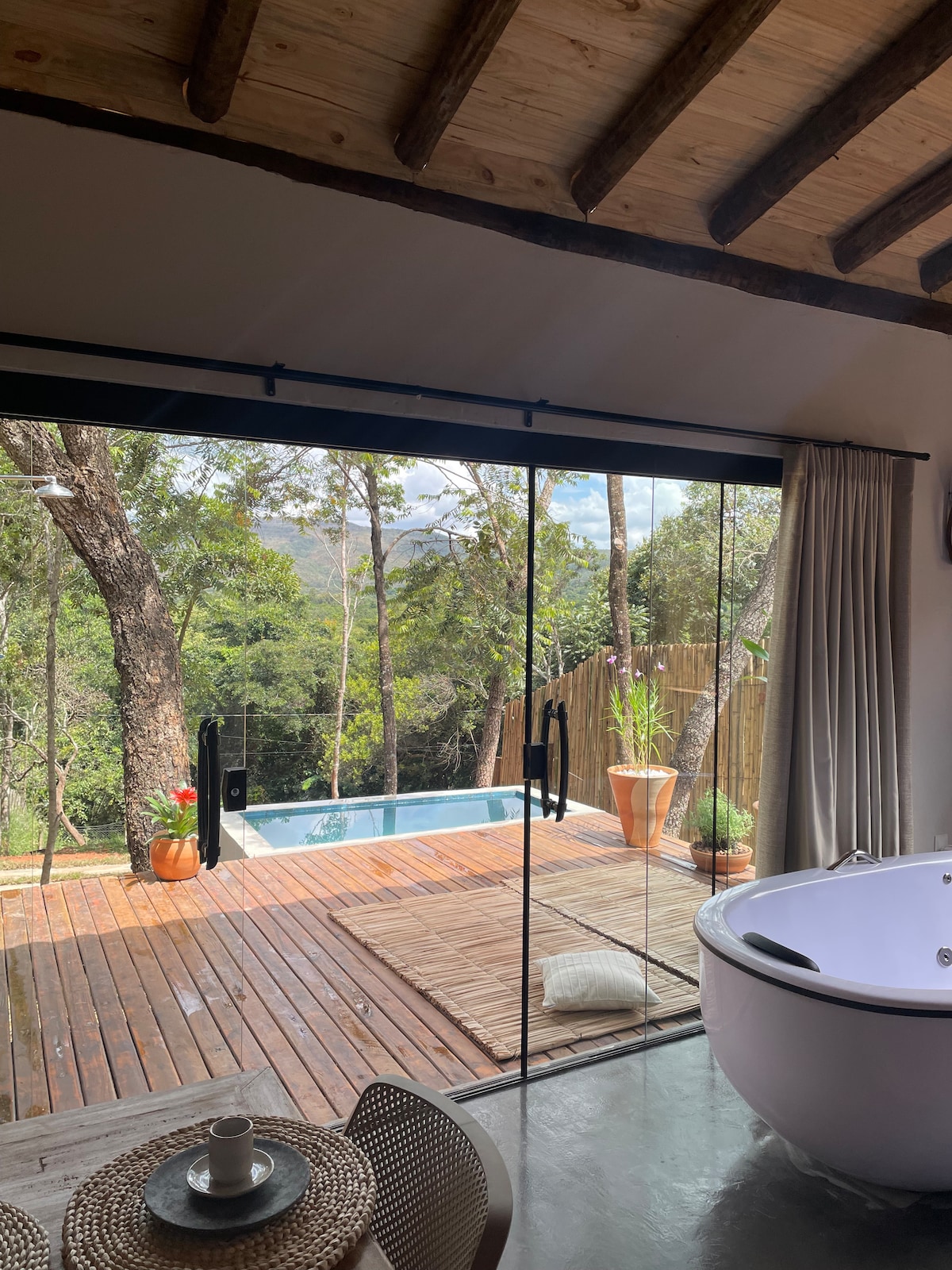 A spacious living area features large glass doors that open to a wooden deck, showcasing a private pool surrounded by greenery. A modern jacuzzi is positioned nearby, and soft natural light filters through sheer curtains, enhancing the serene environment.