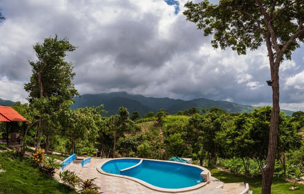 A serene outdoor pool is framed by lush greenery and mountains in the background. The area is surrounded by trees, with a patio nearby featuring comfortable seating. Soft clouds cover the sky, creating a calm atmosphere in this tranquil setting.