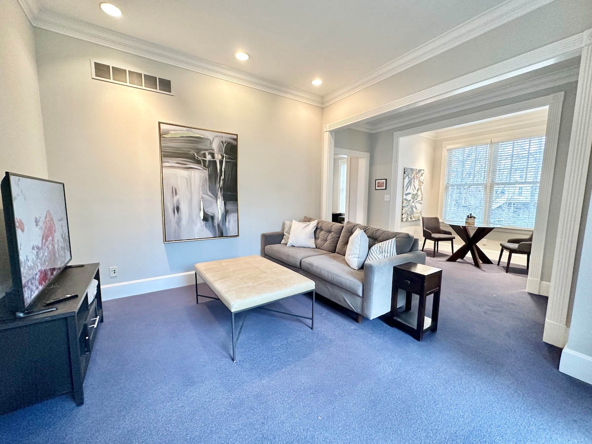 A spacious living area features soft carpet and is furnished with a gray sectional sofa and a coffee table. A large, abstract wall art piece adds visual interest. Natural light enters through windows, illuminating the open layout that leads to the dining area.