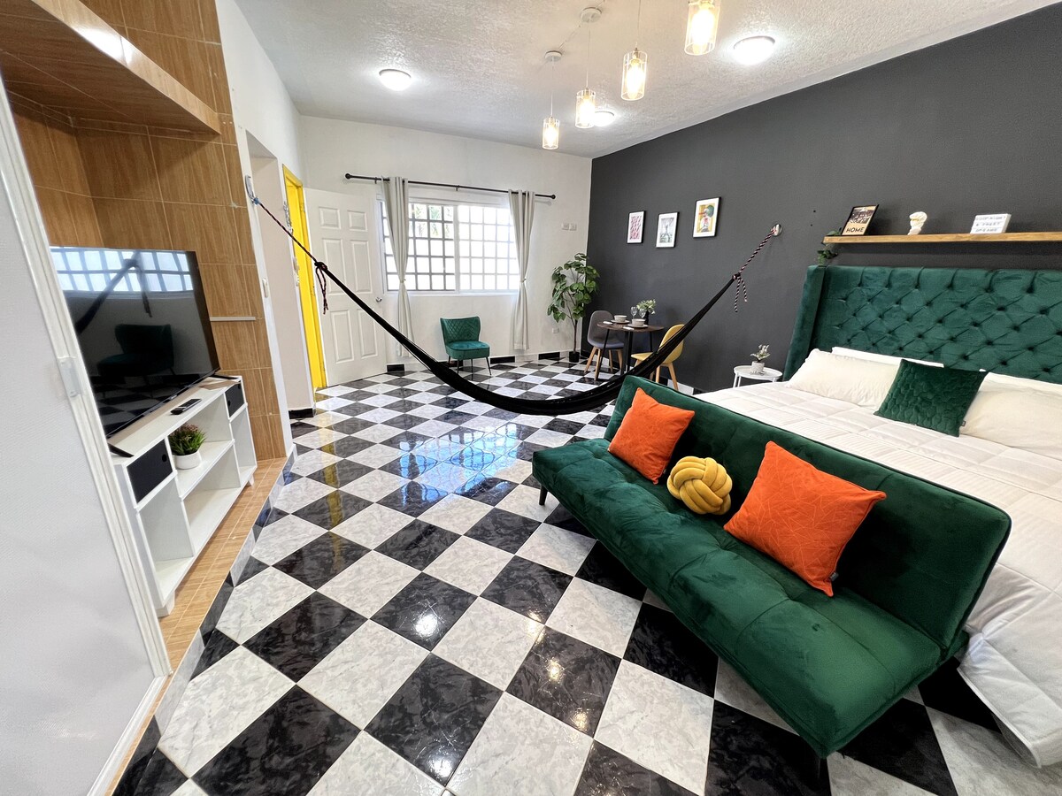A spacious room displays a stylish green sofa adorned with orange cushions and a comfortable king-sized bed. The room features a black and white checkered floor, a wall-mounted television, and a unique hanging hammock for relaxation. Natural light enters through the window, enhancing the ambiance.