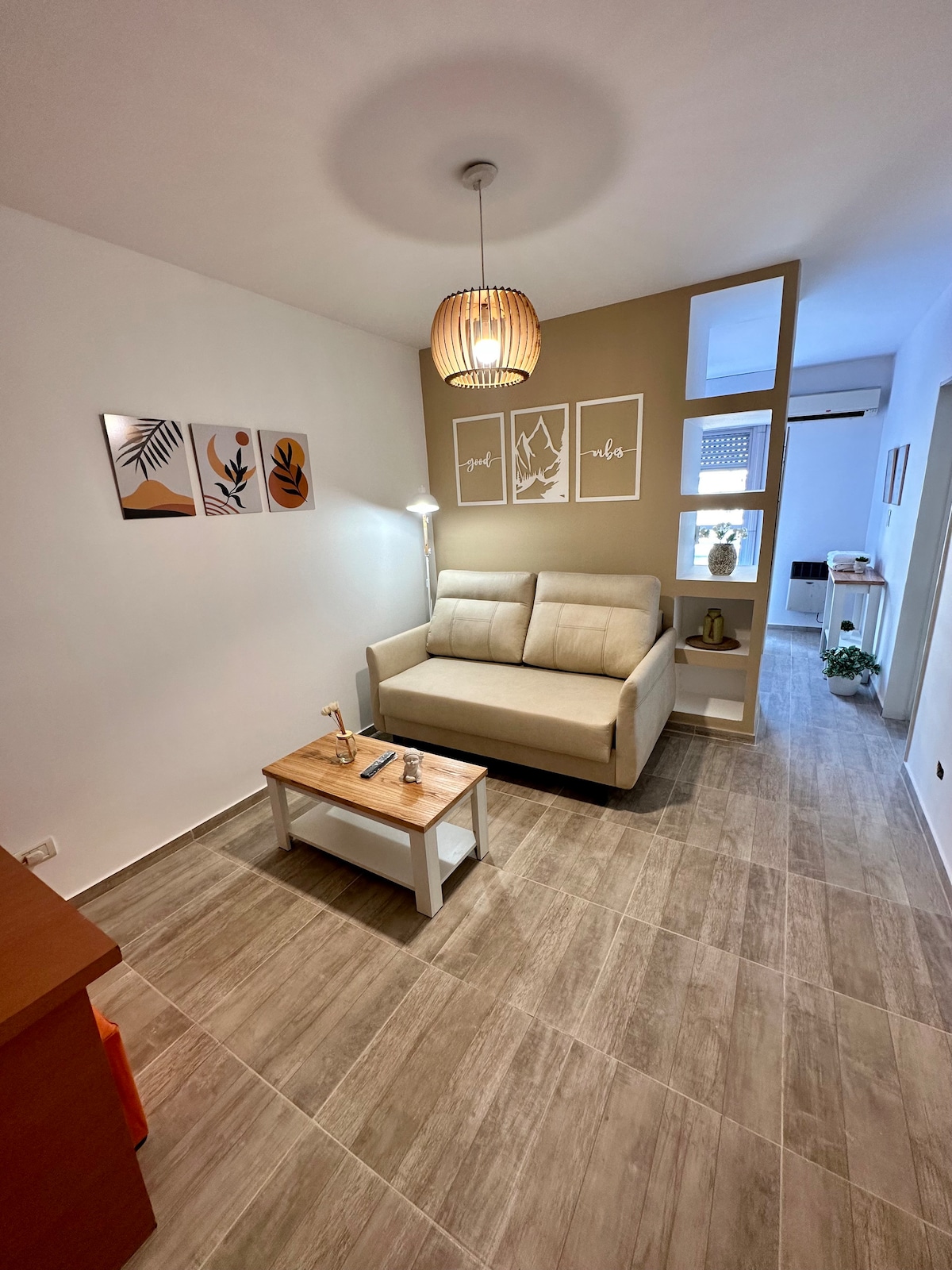 A spacious living area features a comfortable sofa bed, paired with a wooden coffee table. Light beige walls are complemented by framed artwork, and a pendant light hangs from the ceiling. The area is illuminated by natural light from the adjacent space.