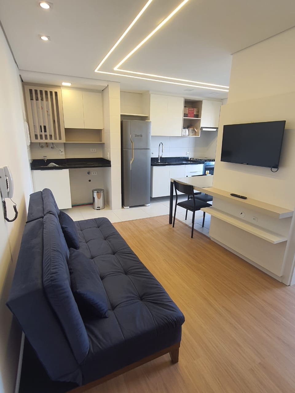 A modern kitchen is equipped with sleek cabinetry, a refrigerator, and a stove. A dining table with black chairs is positioned near a wall-mounted television. Natural light enhances the warm wood flooring, while a comfortable navy blue couch invites relaxation.