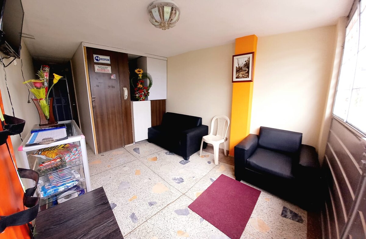 A welcoming reception area is presented with two black sofas and a single white plastic chair. A small dark table is visible, alongside a floral arrangement and a framed picture on the wall. Natural light enters through a nearby window, illuminating the space.