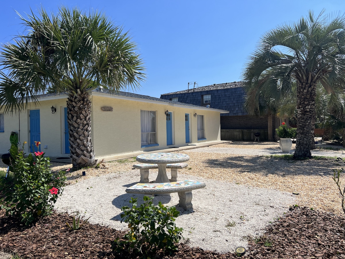 An outdoor area is featured, landscaped with palm trees and rose bushes. A round, mosaic-top table is surrounded by two stone benches, providing a seating space in a gravel-covered yard. The building showcases light-colored walls and blue doors under a bright sky.