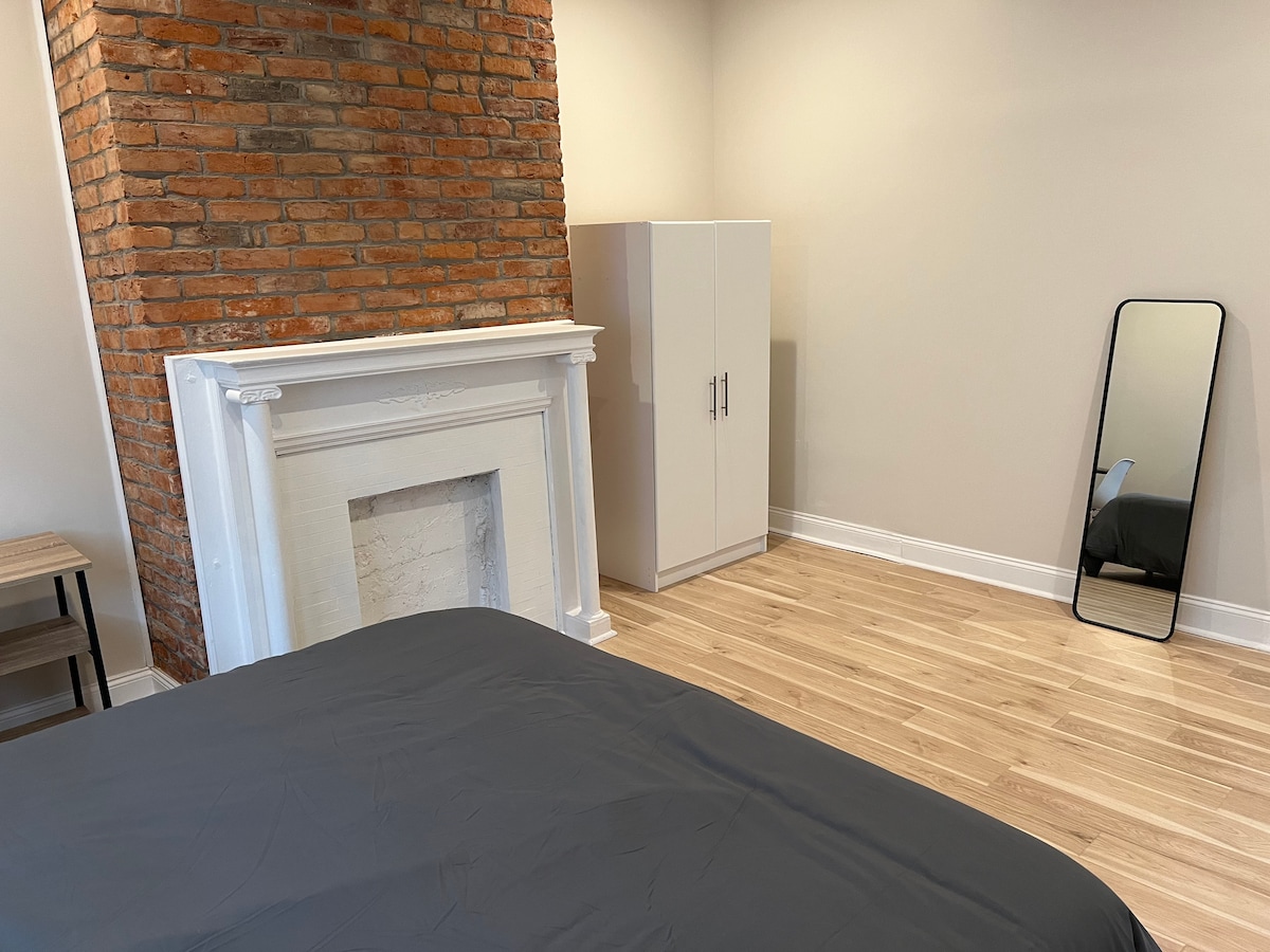 A spacious room features a king-size bed covered with a dark blanket. Textured brick walls and a white fireplace add character. A tall mirror reflects the light, while a closet stands nearby, complemented by wooden flooring enhancing the overall decor.