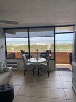 A bright dining area features a round table surrounded by high-backed chairs. Floor-to-ceiling windows provide an unobstructed view of the Atlantic Ocean, with natural light illuminating the space. The tile flooring complements the coastal atmosphere.