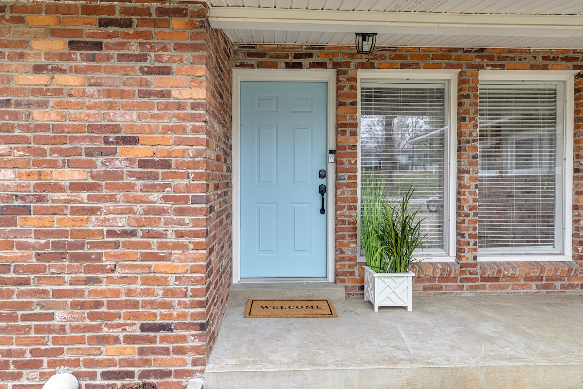 A welcoming entrance features a light blue front door against a backdrop of warm-toned brick. Two large windows, adorned with blinds, allow natural light to brighten the area. A small potted plant adds a touch of greenery beside a 'WELCOME' doormat on the concrete entryway.