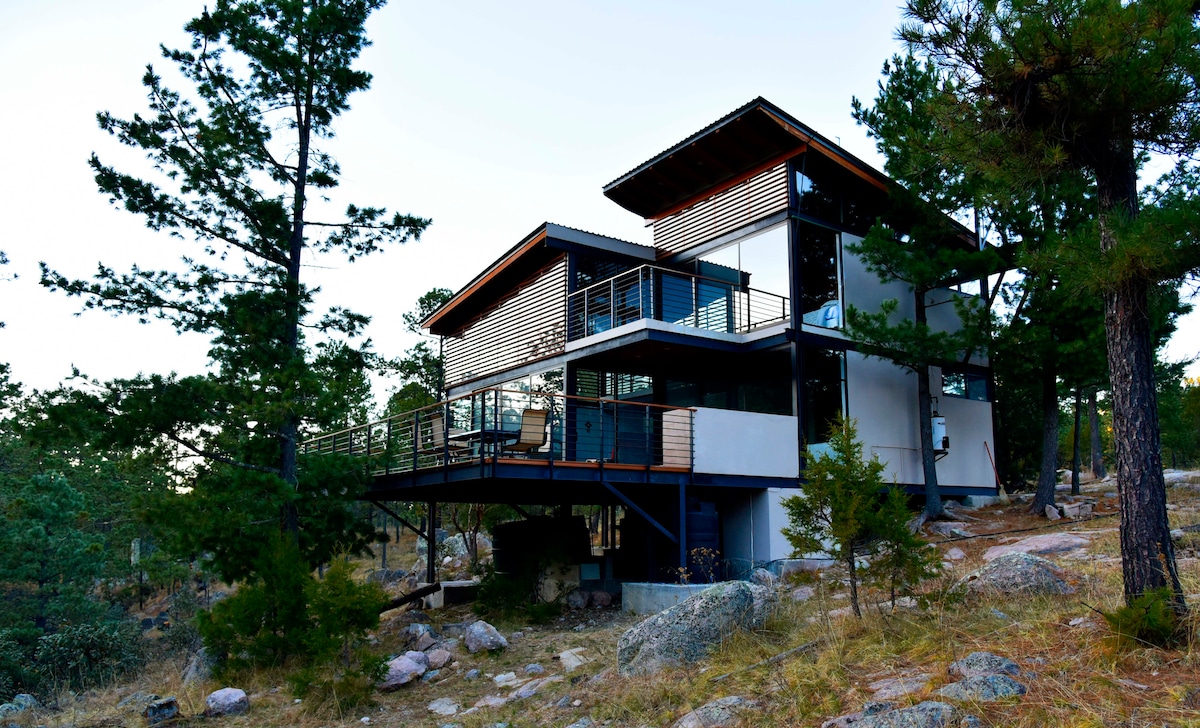 A modern multi-level cabin is situated among trees and rocky terrain. The structure features a blend of wood and metal detailing, with large windows allowing natural light to fill the interior. A spacious balcony overlooks the natural surroundings, enhancing the connection with nature.